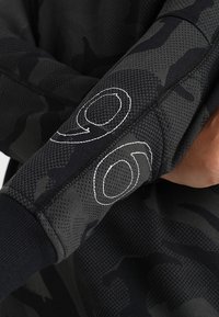 Manche en tissu à motif camouflage gris foncé avec un poignet côtelé noir et les chiffres "90" brodés en blanc sur l'avant-bras.