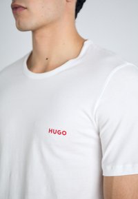 T-shirt en coton blanc à col rond et à manches courtes, avec un petit logo rouge "HUGO" brodé sur la partie gauche de la poitrine.