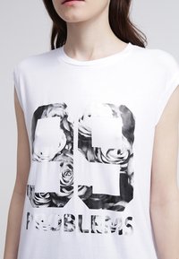 Maglietta bianca senza maniche con un grande "99" e la scritta "PROBLEMS" in nero, decorata con motivi floreali. Tessuto morbido, scollatura rotonda, vestibilità comoda.
