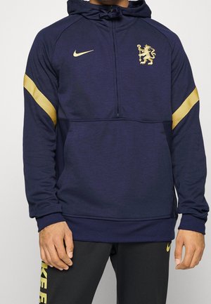 Mann trägt dunkelblauen Nike-Hoodie mit goldenem Löwenemblem und goldenen Streifen an den Ärmeln, kombiniert mit schwarzen Nike-Hosen mit goldenem Logo.
