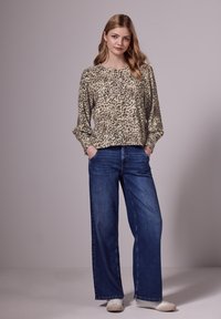 Chemisier beige à manches longues, ample, avec des taches de léopard noires, associé à un pantalon en denim bleu à jambes larges et des baskets blanches.