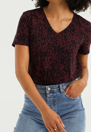 Kobieta w krótkim rękawie, bordowo-czarnym bluzce z dekoltem w serek, włożonej do jasnoniebieskich, wysokich dżinsów z ręką w kieszeni.