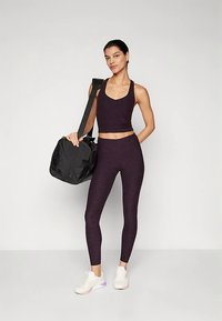 Fialový športový crop top a legíny sú nosené s bielymi teniskami. Čierna športová taška je prehodená cez rameno. Hladká textúra látky.