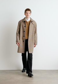 Junger Mann, der geht, trägt einen beigefarbenen karierten Mantel, einen braunen Pullover über einem weißen Hemd, schwarze Hosen und schwarze Schuhe vor einem schlichten weißen Hintergrund.