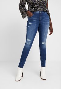 Jeans skinny azul con un diseño desgastado y rasgado, con desgarros en las rodillas y un estilo clásico de cinco bolsillos. Llevados con botas de tacón blancas.