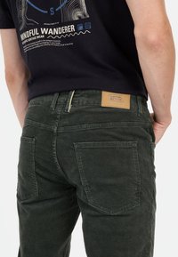 Pantalons en velours côtelé vert foncé avec une étiquette à la taille. Deux poches arrière avec un détail de couture visible, matériau texturé et coupe slim.