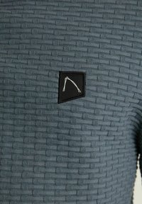 Textured teal sweater featuring a woven pattern, with a black label displaying a white design. The fabric is soft with a slightly raised texture.