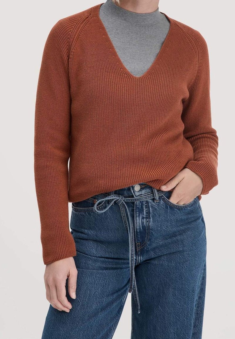 Personne portant un pull col V de couleur rouille par-dessus un col roulé gris, associé à un jean en denim bleu avec une ceinture nouée.