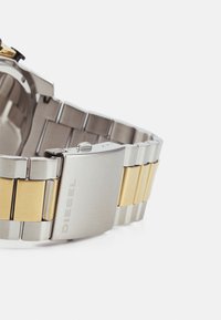 Orologio in metallo argento e oro con finitura spazzolata, dotato di un bracciale bicolore e una chiusura quadrata con il nome del marchio "DIESEL" inciso.
