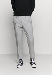 Osoba v šedých slim-fit kalhotách s řetízkem, černých vysokých teniskách a černém dlouhém rukávu, stojící na světlém pozadí.