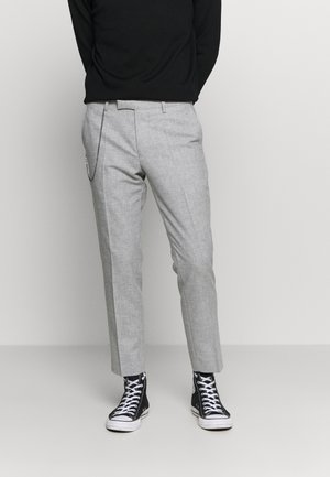 Personne portant un pantalon gris slim, avec un accessoire chaîne, des baskets montantes noires et un haut à manches longues noir, se tenant sur un fond clair.