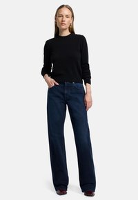 Pull nero a maglia con maniche lunghe abbinato a jeans scuri a vita alta e gamba larga. Il modello si trova su uno sfondo neutro.