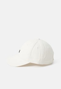 Hvitt bomullsbasker med buet skjerm og svart logo-detall på forsiden. Har ventilasjons hull og justerbar stropp bak.