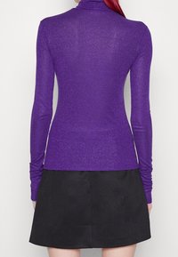 Lila långärmad topp gjord av ett glittrande, stretchigt material, med högt krage och figurnära design, som bärs med en svart kjol.