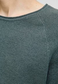 Tricot de culoare verde închis, cu o suprafață texturată, având un guler larg și mâneci raglan, prezentând detalii ale modelului de tricot.