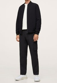 Veste bomber noire avec fermeture éclair, t-shirt blanc à col rond en dessous, pantalon cargo noir et baskets blanches. Tissu lisse, coupe moderne.