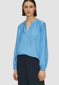 Lichtblauw, langmouw blouse met een V-hals, flatterende plooien bij de schouders en een gestructureerde stof, gecombineerd met donkere broeken.