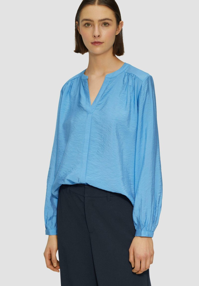 Lichtblauw, langmouw blouse met een V-hals, flatterende plooien bij de schouders en een gestructureerde stof, gecombineerd met donkere broeken.