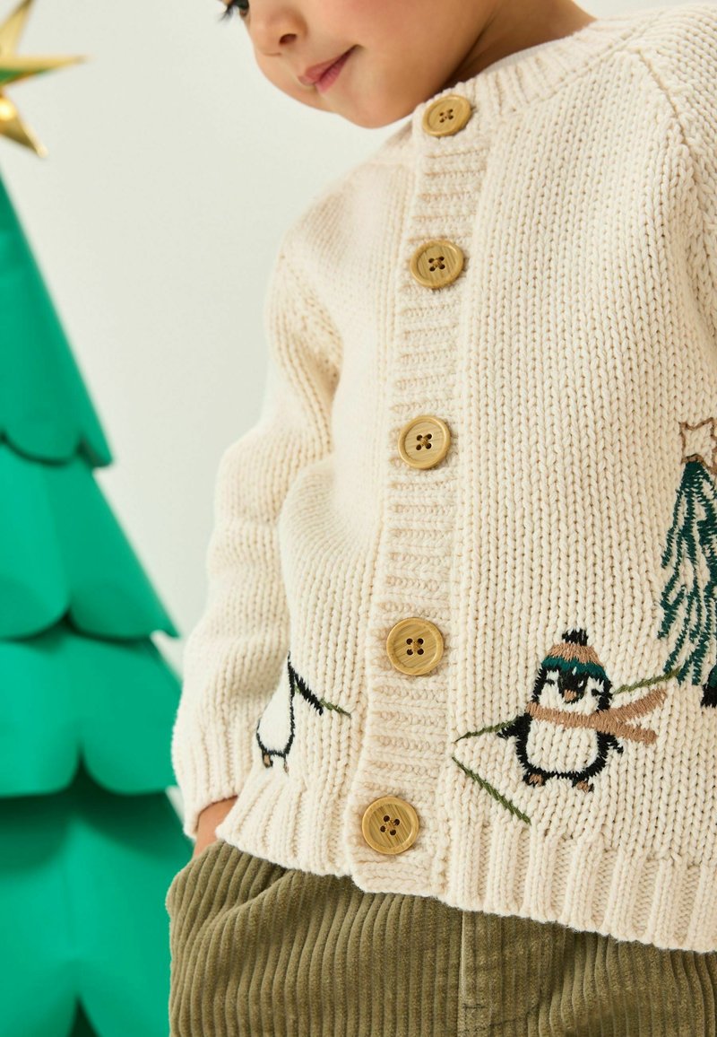 Cremefarbener Strickcardigan mit Holzknöpfen, bestickten Pinguinen und Bäumen, mit geripptem Saum, kombiniert mit olivgrünen Cordhosen.
