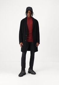 Cappotto nero con grandi bottoni, indossato sopra un maglione rosso. Berretto nero con logo, jeans scuri e sneakers nere texture.