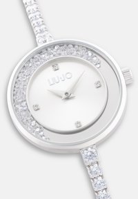 Silberne Uhr mit einem glatten Ziffernblatt, Kristallverzierungen und einem runden Gehäuse. Verfügt über ein mit Edelsteinen besetztes Armband und minimalistische Stundenmarkierungen.
