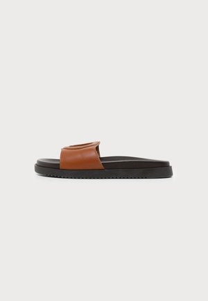Une sandale mule en cuir marron avec une semelle noire, un talon plat et une large bride sur le dessus, vue de profil sur fond blanc.