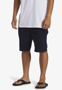 Bermudas de algodón azul marino de corte recto, hasta la rodilla, con bolsillos delanteros, combinadas con chanclas negras y una camiseta blanca de cuello redondo.