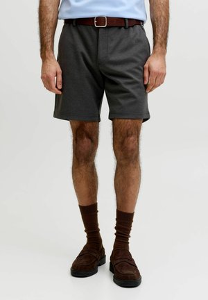 Uomo che indossa pantaloncini casual grigio scuro, cintura marrone, calzini marroni e mocassini marroni, in piedi davanti a uno sfondo grigio chiaro.