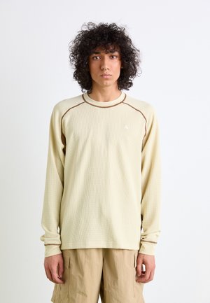 Long-sleeve, light beige shirt with a textured fabric, raglan sleeves, contrasting brown stitching, and a small triangular logo on the chest.