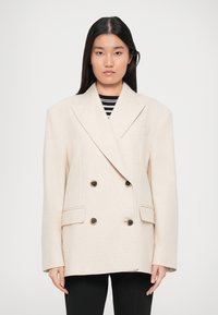 Woman with long black hair wearing a double-breasted cream blazer over a black and white striped shirt and black pants, standing against white background.