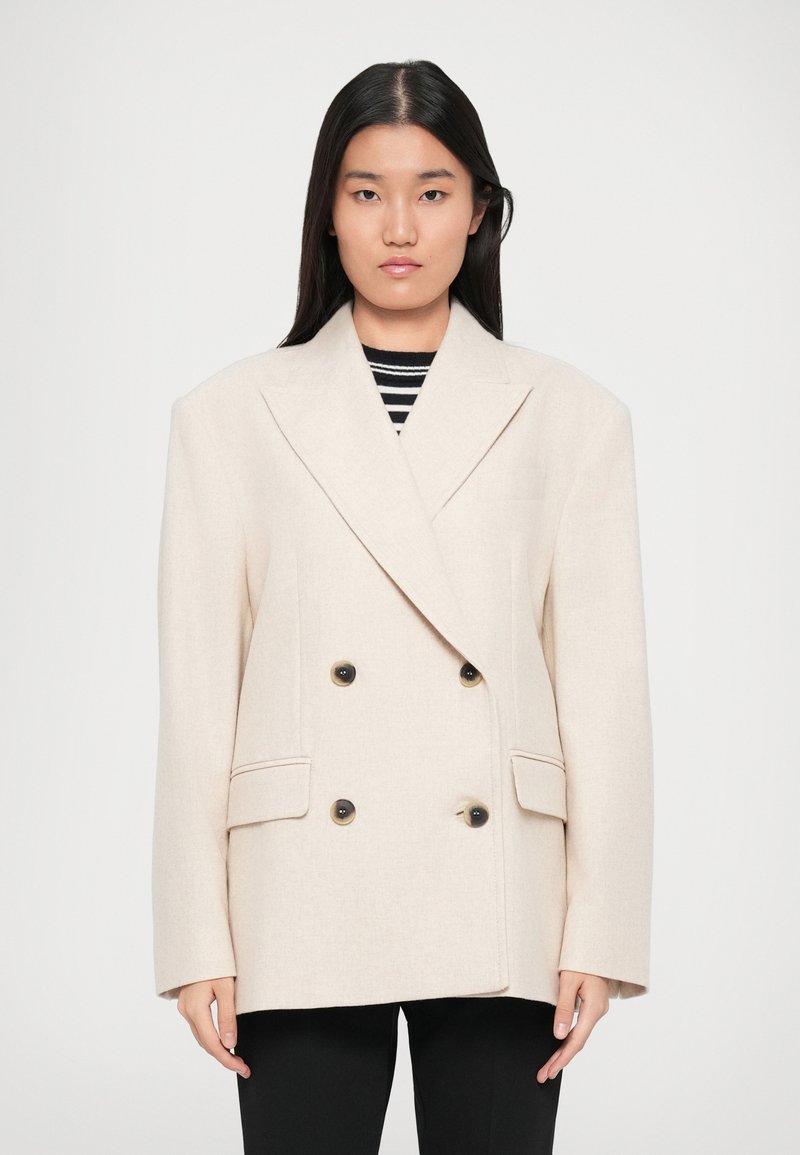 Woman with long black hair wearing a double-breasted cream blazer over a black and white striped shirt and black pants, standing against white background.