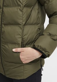 Olivový zelený páperový kabát so zipsom, obsahujúci bočné vrecko a čierne lemovanie na manžetách. Hladká textúra a prešívaný dizajn.
