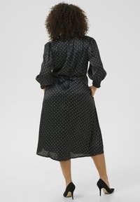 Zwarte jurk met stippen, gemaakt van gladde stof, met een ceintuur in de taille, gathering op de mouwen en een hem die tot de knie reikt. Gestyled met zwarte hakken.
