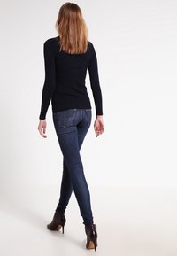 Kvinna med långt hår iklädd mörka skinny jeans, svart långärmad topp och bruna högklackade ankelstövlar som går bortåt mot en enfärgad vit bakgrund.