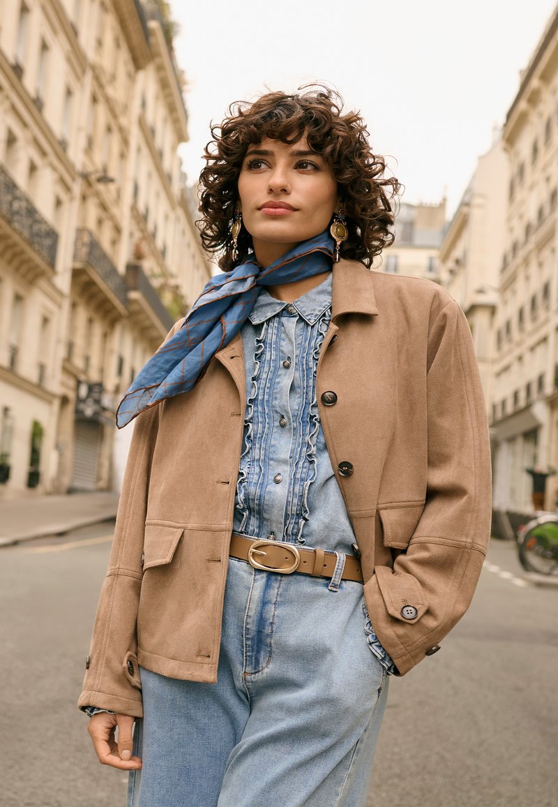 Jachetă maro din piele intoarsă, cămașă denim cu volane, salopetă denim deschis la culoare, eșarfă albastră cu accente portocalii, cercei cerc din aur, cadru urban casual.