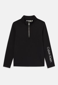 Crna dugom rukavu majica s polu-zipom i visokim ovratnikom, izrađena od mekanog materijala. Ima bijeli natpis "Calvin Klein" na desnoj rukavnoj strani.