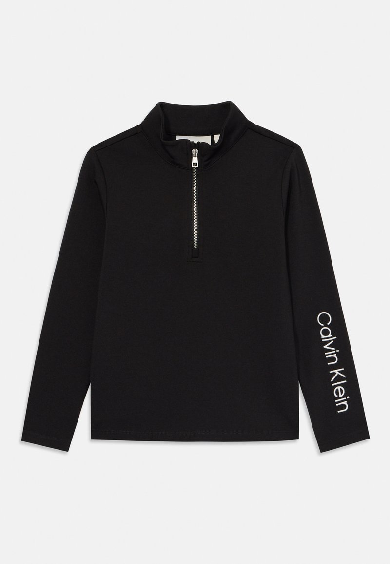 Crna dugom rukavu majica s polu-zipom i visokim ovratnikom, izrađena od mekanog materijala. Ima bijeli natpis "Calvin Klein" na desnoj rukavnoj strani.