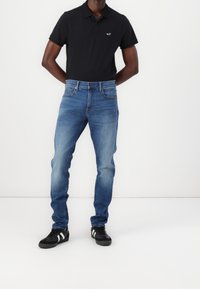 Camisa polo azul oscuro con logo, jeans azules claros con textura desgastada y zapatillas negras con rayas blancas. Conjunto casual exhibido en un modelo.