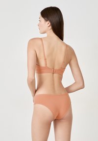 Biustonosz i majtki w zestawie wykonane z miękkiego, elastycznego materiału. Oba elementy są w jasnobrązowym kolorze, mają regulowane ramiączka oraz zapięcie na klamrę w biustonoszu.