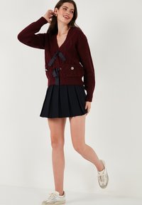 Burgundy knitted cardigan with a V-neck, featuring two pockets with bows, paired with a pleated navy skirt and light sneakers.