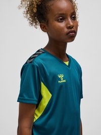 Fille aux cheveux bouclés portant un maillot de sport bleu sarcelle avec des accents jaunes et le logo hummel, regardant légèrement sur le côté sur un fond uni.