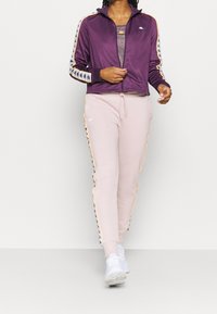 Veste zippée violette avec des accents rayés blancs et noirs, associée à un jogging rose pâle avec des logos latéraux et un cordon élastique.