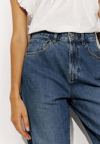 Jean bleu taille haute avec une texture délavée, quatre poches, des coutures contrastantes et une fermeture à bouton à la taille.