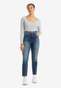 Γυναίκα που στέκεται με το χέρι στην τσέπη, φορώντας γκρι μακρυμάνικο πουκάμισο με κουμπιά, ψηλόμεσα μπλε τζιν, λευκές κάλτσες και λευκά sneakers.