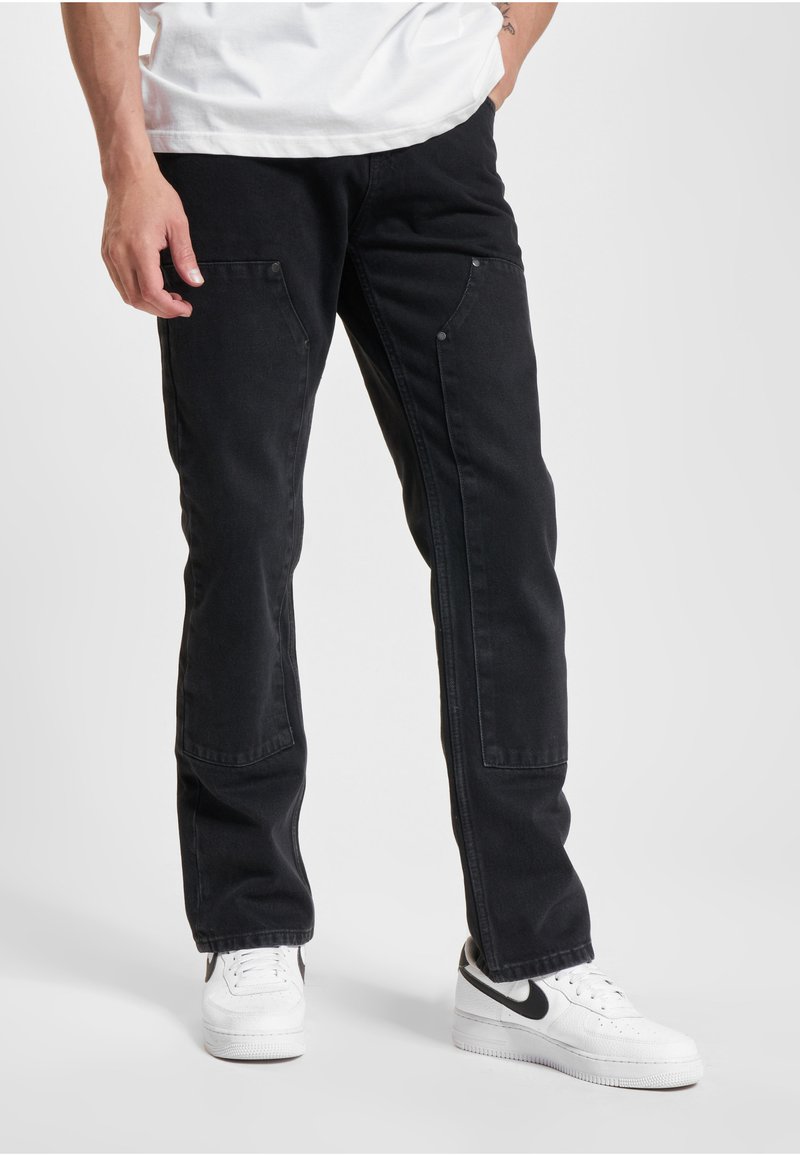 Mies, jolla on mustat monitaskuiset cargo-housut ja valkoiset lenkkarit mustalla swoosh-logolla, seisoo yksivärisen taustan edessä.