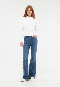 Biały sweter w warkoczyki z wysokim dekoltem, w połączeniu z niebieskimi spodniami dżinsowymi w stylu dzwonów i białymi butami sportowymi na jasnym tle.