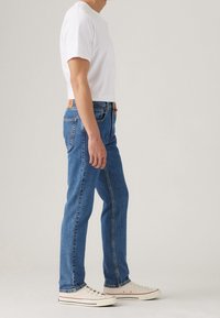 Modré džínové kalhoty s rovnými nohavicemi, pětikapsový styl, vysoký pas s oranžovou štítkem. Kombinovány s jednobarevným bílým tričkem a bílými teniskami.