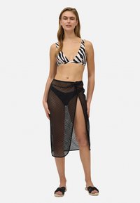 Cubierta de playa de malla negra con un detalle anudado, combinada con un top de bikini de rayas de cebra. Diseño abierto con una textura transparente.