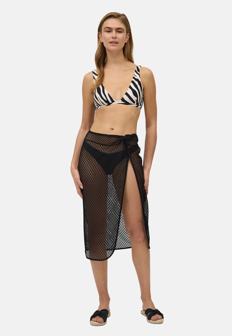 Cubierta de playa de malla negra con un detalle anudado, combinada con un top de bikini de rayas de cebra. Diseño abierto con una textura transparente.