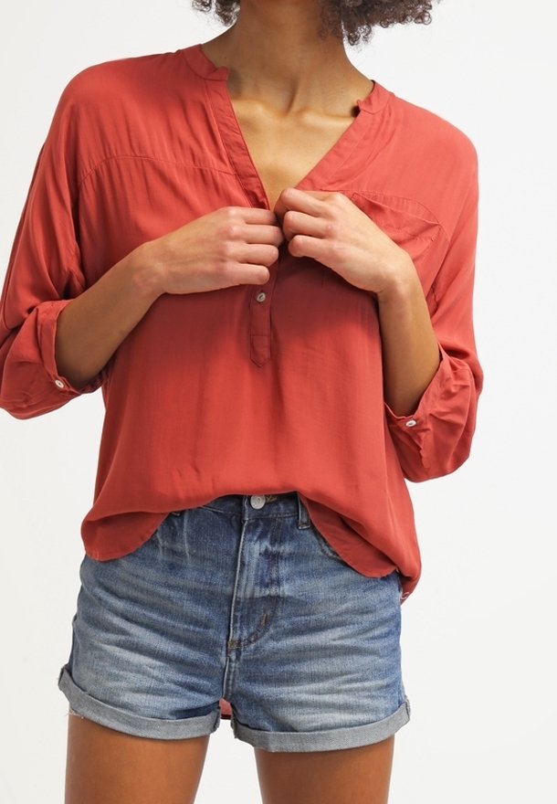 Personne portant un chemisier rouge ample à manches longues et un short en denim bleu retroussé, ajustant les boutons du chemisier avec les deux mains.
